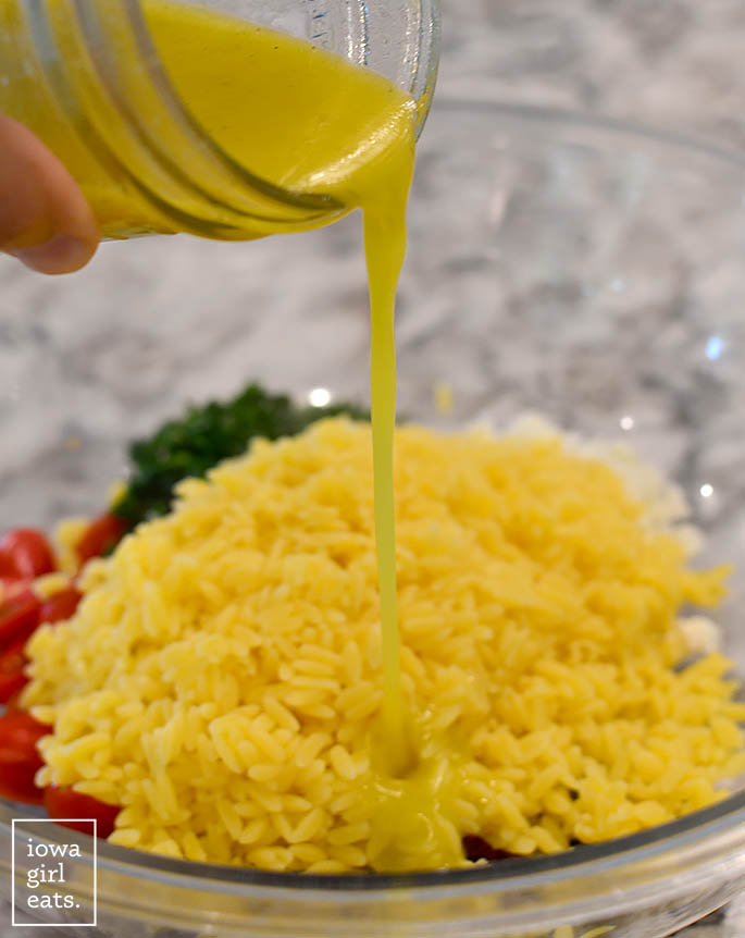lemon dressing being poured over the ingredients for mediterranean orzo salad