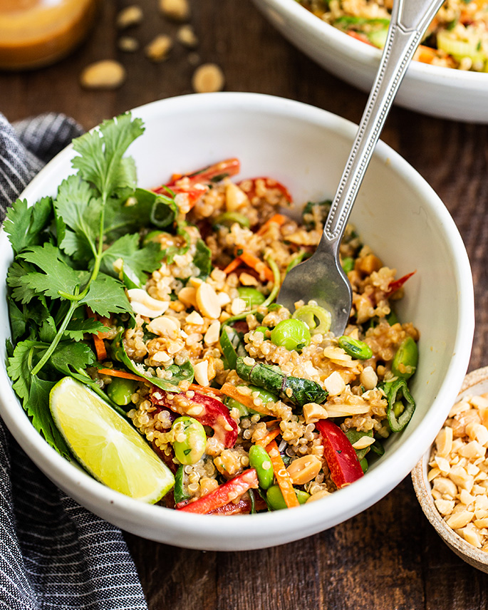 a fork in a bowl of thai quinoa salad with fresh herbs and lime wedges